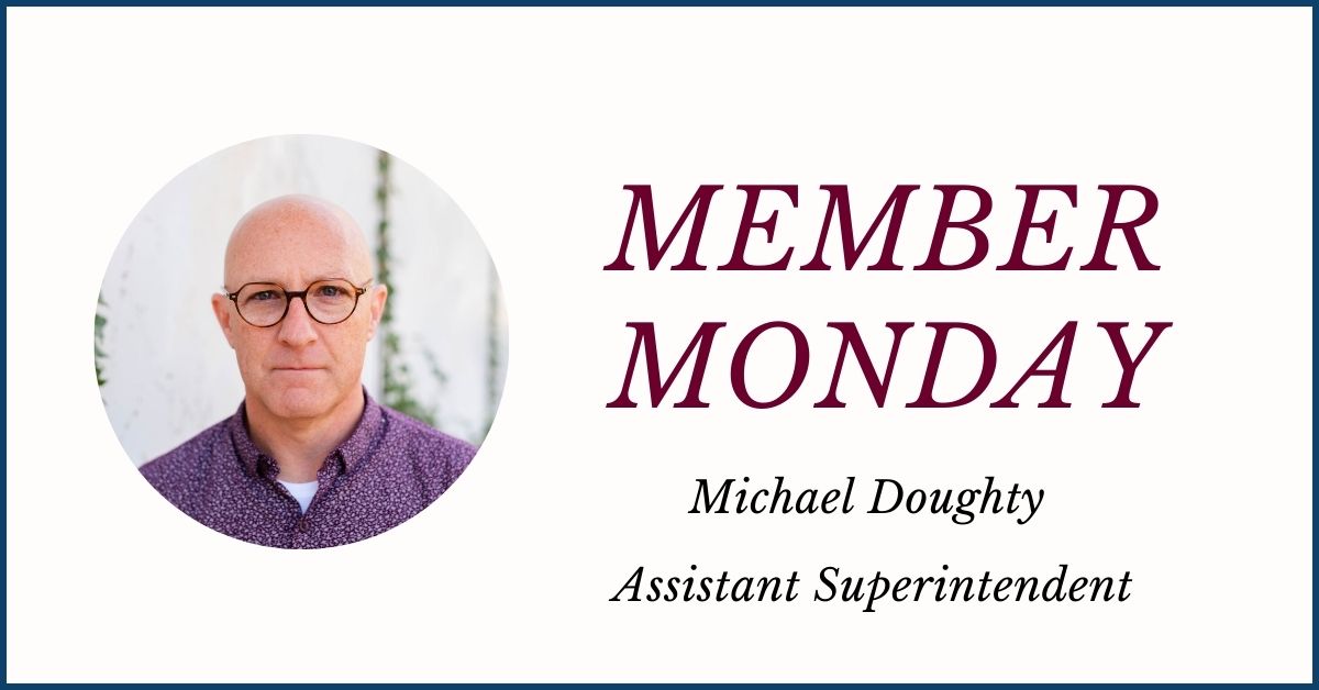 An icon featuring a circular photo at left of Michael Doughty, who wears round tortoise shell eyeglasses, a blue, open-collared dress shirt and is looking at and smiling for the camera. The words “Member Monday Michael Doughty, Assistant Superintendent” are displayed at the right.