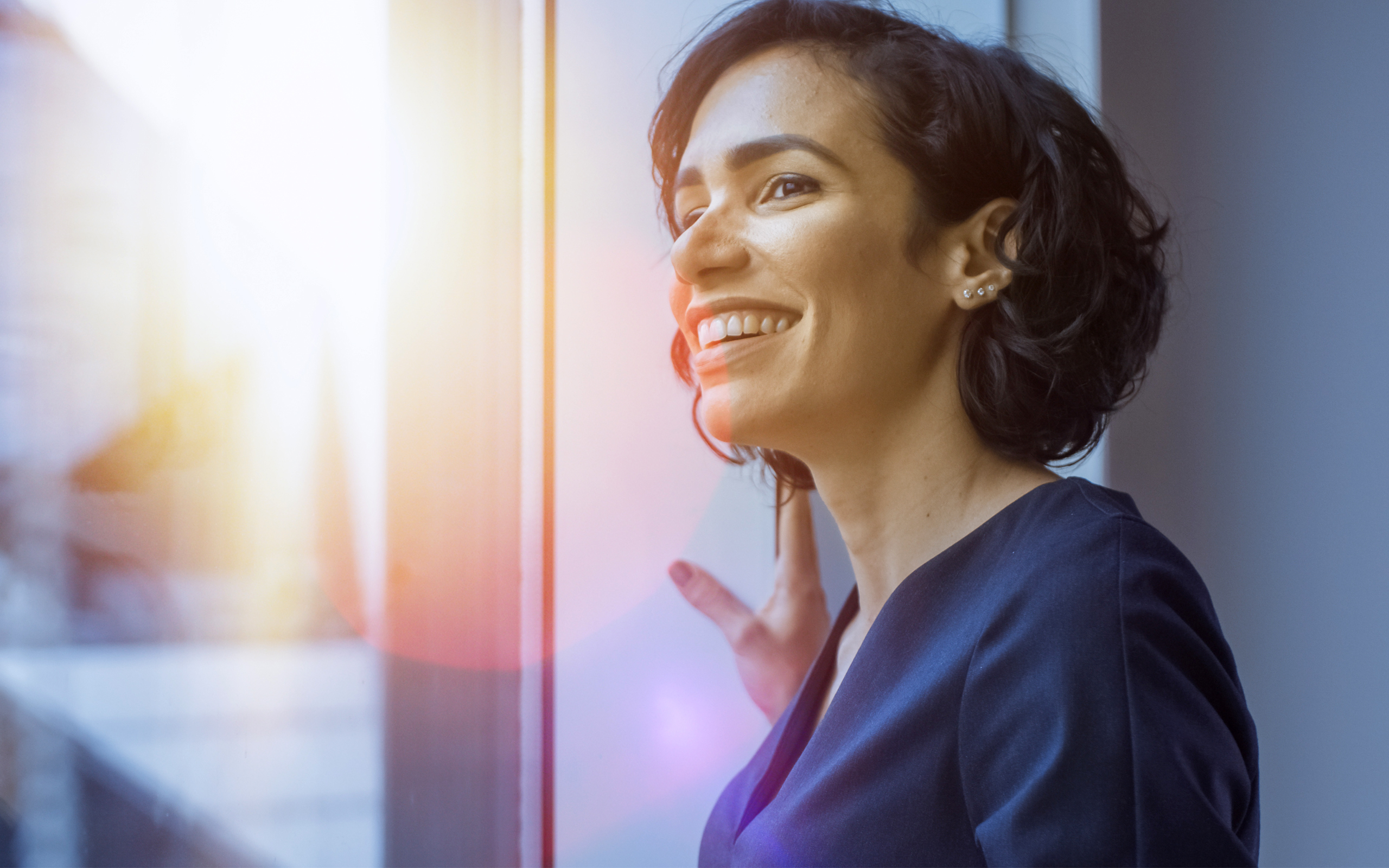 Smiling, confident woman, looking out window of office with sunlight outside