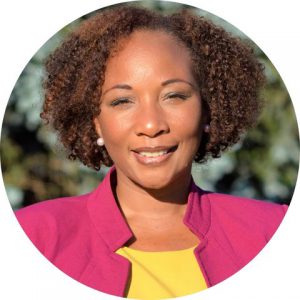 Dr. Monique Darrisaw-Akil, who has short brown curly hair, is wearing a yellow blouse and pink jacket and stands in front of a lush green background, looks at and smiles for the camera.