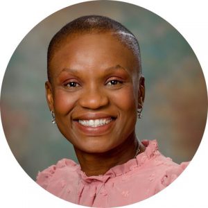 Nerlande Anselme, who has short dark hair, is wearing a pink blouse and stands in front of a green background, looks at and smiles for the camera.