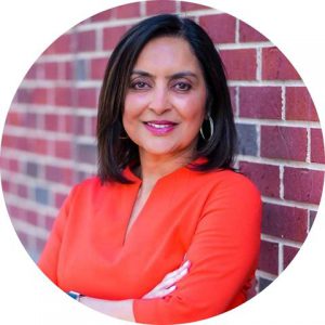 Sujata Chaudhry, who has mid-length dark hair, is wearing an orange blazer and leaning against a brick wall with her arms crossed. 
