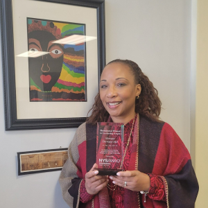 Dr. Monique Darrisaw-Akil stands in front of a framed painting. Dr. Akil-Darrisaw is holding up a crystal award.